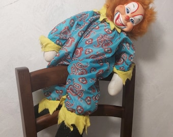 May include: A blue and red clown doll with a yellow and white patterned outfit sits on a wooden chair. The doll has a red wig and a painted-on smile.