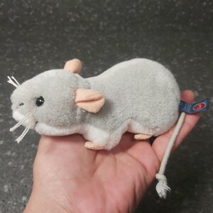 Op de afbeelding: Een zachtgrijs rattenknuffel met roze oren en voeten, en witte snorharen. Het speelgoed heeft een lange, witte touwstaart en een blauw label. De rat wordt in een hand gehouden.