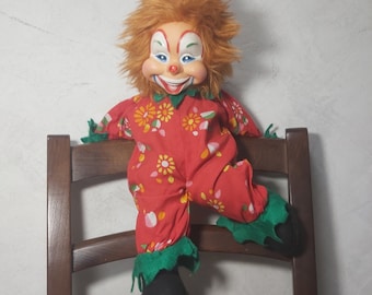 May include: A red and white clown doll with a red and green patterned jumpsuit sits on a wooden chair. The clown has a red nose, white face paint, and a big smile.