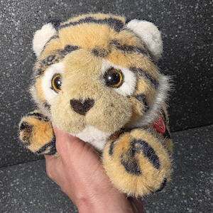 Op de afbeelding: Een pluche tijgerknuffel met goudbruine vacht met zwarte strepen, witte accenten op de oren en snuit, en grote bruine ogen. Het knuffeldier heeft een kleine donkere neus en een rond rood label bevestigd aan de zijkant van het lichaam.