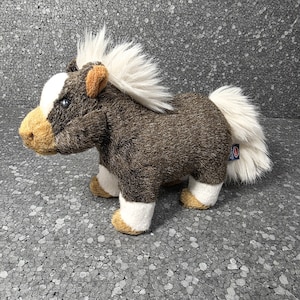 Op de afbeelding: Een pluchen pony met een bruin lichaam, witte manen en staart, en bruine accenten op de snuit, oren en hoeven. Het speelgoed heeft een witte vlek op het voorhoofd en een klein label op de achterkant.