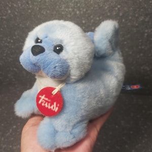 Op de afbeelding: Een blauw pluche zeehondenspeelgoed met een witte buik en kop. De zeehond heeft zwarte ogen en een zwarte neus. Een rood rond label met het woord "Trudi" is aan de zeehond bevestigd.