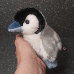 Op de afbeelding: Een pluche pinguïn met een zwarte kop, wit gezicht en grijze lichaam. De pinguïn heeft een blauwe snavel en voeten. Het speelgoed wordt in een hand gehouden tegen een donkere achtergrond. Een klein label is zichtbaar aan de zijkant.