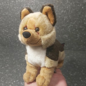 Op de afbeelding: Een bruin en wit pluche hondenspeelgoed met een gestikte litteken op zijn gezicht. Het speelgoed heeft bruin haar op zijn rug en wit haar op zijn buik en poten.