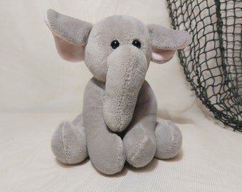 May include: A gray plush elephant toy with pink ears and black eyes. The elephant is sitting with its legs crossed and its trunk curled up.