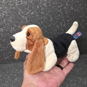 Op de afbeelding: Een pluche basset hound-speeltje met een witte, bruine en zwarte vacht, lange zachte oren, een zwarte neus en donkere ogen. Het knuffeldier heeft een klein blauw label op de rug met de tekst 'trudi' in witte letters gedrukt.