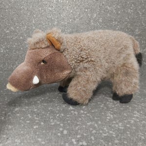 Peut inclure: Un jouet en peluche de sanglier brun et gris avec une défense blanche. Le jouet a une texture pelucheuse et des yeux noirs.