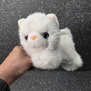 Op de afbeelding: Een kleine, pluizige witte knuffel in de vorm van een kitten. Het speelgoed heeft zachte, gestructureerde witte vacht, twee grote, ronde, donkere plastic ogen en een kleine, lichtgekleurde, driehoekige neus. De oren zijn puntig en rechtopstaand, en het algehele uiterlijk is dat van een jonge, langharige kat met een rond gezicht en een dichte, zachte vacht.