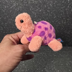 Puede incluir: Un pequeño juguete de tortuga de peluche con cabeza y extremidades de color melocotón, que presenta un caparazón rosa brillante adornado con manchas moradas. La tortuga tiene ojos de plástico negro, una etiqueta blanca y una etiqueta azul.