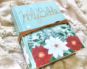 May include: A light blue Holy Bible with a floral design painted on the cover. The flowers are white, red, and green. A brown leather strap is wrapped around the book.