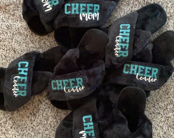 May include: Six pairs of black fuzzy slippers with different turquoise blue glitter text. The text on the slippers reads "Cheer Mom" and "Cheer Leader".