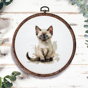 May include: A cross-stitch embroidery of a Siamese kitten with blue eyes, set in a wooden hoop. The kitten is light brown with darker brown markings. The background is white with brown watercolor-like splatters.