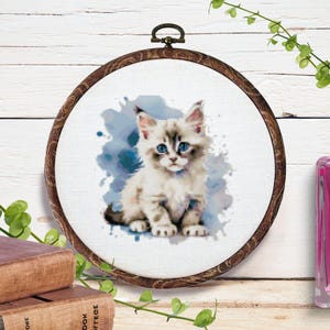 May include: A watercolor-style embroidery hoop featuring a fluffy kitten with blue eyes and gray markings. The cat is set against a blue watercolor splash. The wooden hoop has a metal loop for hanging. The artwork is displayed on a white wooden background.