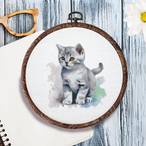 May include: A round, wooden embroidery hoop featuring a watercolor painting of a gray and white kitten with yellow eyes. The artwork is set against a white background. The hoop is on a light blue wooden surface.