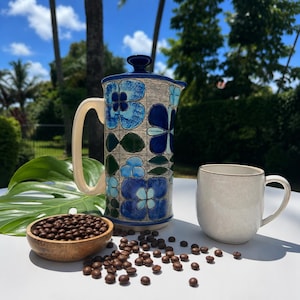 May include: A ceramic French press featuring a blue and white floral pattern, a cream-colored handle, and a matching lid. Beside it sits a plain cream-colored ceramic mug and a small wooden bowl filled with dark roasted coffee beans.