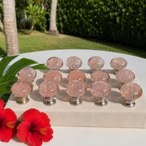 May include: A collection of fourteen round cabinet knobs made of clear glass with a pink tint and internal bubble textures, each featuring a metallic silver-toned base, arranged in rows on a rectangular off-white fabric display block.