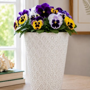 May include: A white ceramic planter with a raised, repeating arch pattern containing a vibrant arrangement of purple, yellow, and white pansy flowers with green leaves. The planter has a tapered, cylindrical shape and a glossy finish.