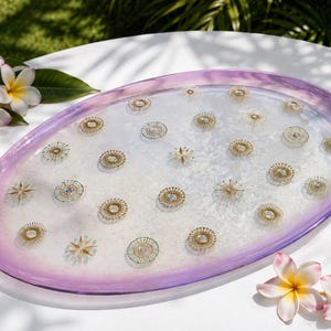 May include: An oval-shaped decorative tray with a translucent white center and a gradient purple rim. The surface is embedded with numerous small, circular gold-toned metallic accents and star-shaped motifs arranged across the clear base.