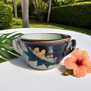 May include: A ceramic bowl with two side handles, featuring a dark blue and cream glaze with a textured, orange-speckled floral pattern on the exterior. The interior is a smooth, light cream color, and the rim is finished with a dark brown glaze.