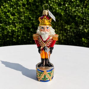 May include: A glass nutcracker ornament featuring a gold crown, white beard, and red uniform with gold accents. The figure stands on a cylindrical base decorated with green holly leaves and gold trim, topped with a silver hanging loop and a small tag.