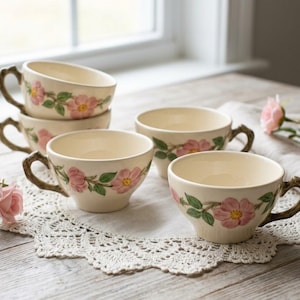 May include: A set of six cream-colored teacups with brown handles, each adorned with pink rose and green leaf designs. The cups are arranged on a white lace doily, perfect for a tea party.