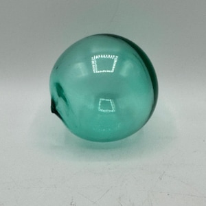 May include: A teal-colored, spherical glass float. The float has a smooth, rounded shape with a small, dark protrusion on one side. The glass appears translucent, reflecting light and creating highlights. The object is likely used for decoration or as a nautical element.
