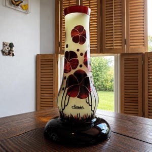 May include: A tall, decorative vase with a wide, black base and a cream-colored body. The vase features large, red floral designs and a dark red rim. The word "Charder" is written on the vase. The vase is displayed on a wooden table.