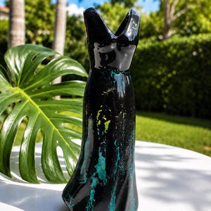 May include: A glossy black ceramic vase shaped like a sleeveless dress with a V-neckline. The surface features a textured, marbled pattern with streaks of vibrant turquoise and bright yellow paint accents running vertically down the flared base.