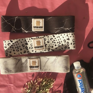 Op de afbeelding: Drie weerstandsbanden met marmer- en luipaardpatronen. Elke band heeft een wit vierkant label met de tekst "POWER HOUSE PERFORMANCE FITNESS". Een fles water en een klein boeket droogbloemen zijn ook aanwezig.