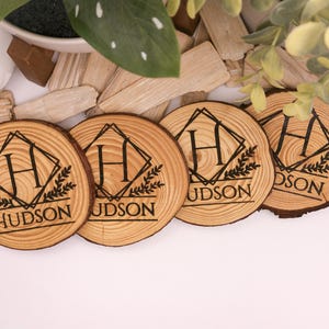 May include: Four wooden coasters with a natural wood grain. Each coaster features a black geometric design with the letter "H" and the name "HUDSON". The coasters are arranged on a white surface with wood chips and greenery.