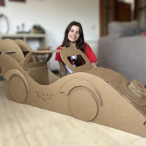 May include: A brown cardboard Batmobile-style car. The car features circular wheels, a bat symbol, and a detailed rear section. The car is designed for children. The car's dimensions are approximately 150 cm in length.