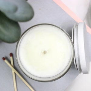 May include: A white candle in a clear glass jar with a gray lid. The candle is unlit and has a small indentation in the center.