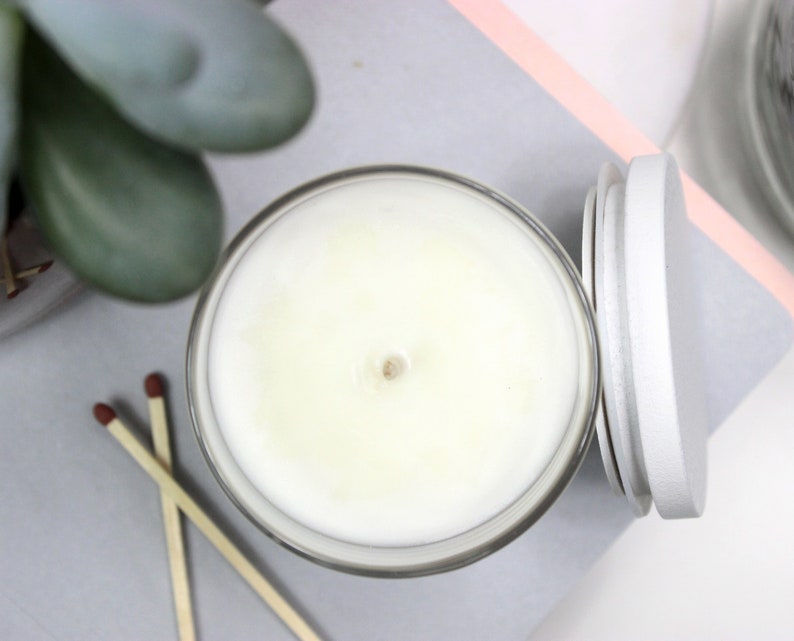 May include: A white candle in a clear glass jar with a gray lid. The candle is unlit and has a small indentation in the center.