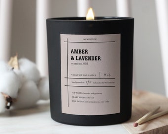 May include: Black soy wax candle with a pink label that reads "WICKITSTUDIO", "AMBER & LAVENDER", "scent no. 003", "VEGAN SOY WAX CANDLE", "30 cl", "hand poured on 7/11 in London by WickitStudio", "TOP NOTES: lavender and geranium", "HEART NOTES: softwoods", "BASE NOTES: amber, frankincense and tonka".