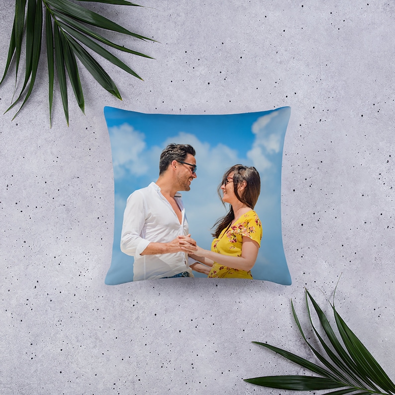 May include: A square throw pillow featuring a photograph of a couple gazing at each other against a blue sky with white clouds. The man is wearing a white shirt and sunglasses, and the woman is wearing a yellow dress and sunglasses.