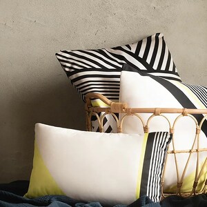 May include: Three decorative cushions with black and white geometric patterns and a yellow accent. The cushions are arranged in a wicker basket on a bed with a dark blue throw blanket.