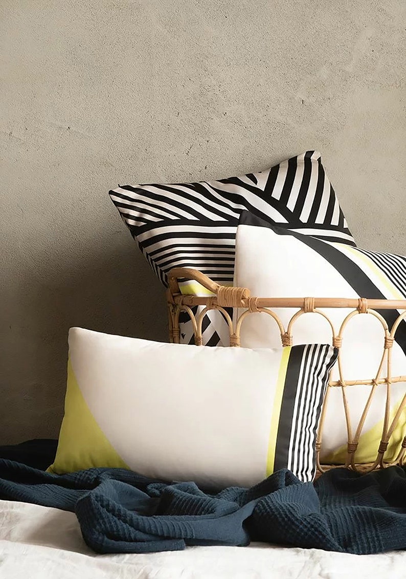 May include: Three decorative cushions with black and white geometric patterns and a yellow accent. The cushions are arranged in a wicker basket on a bed with a dark blue throw blanket.