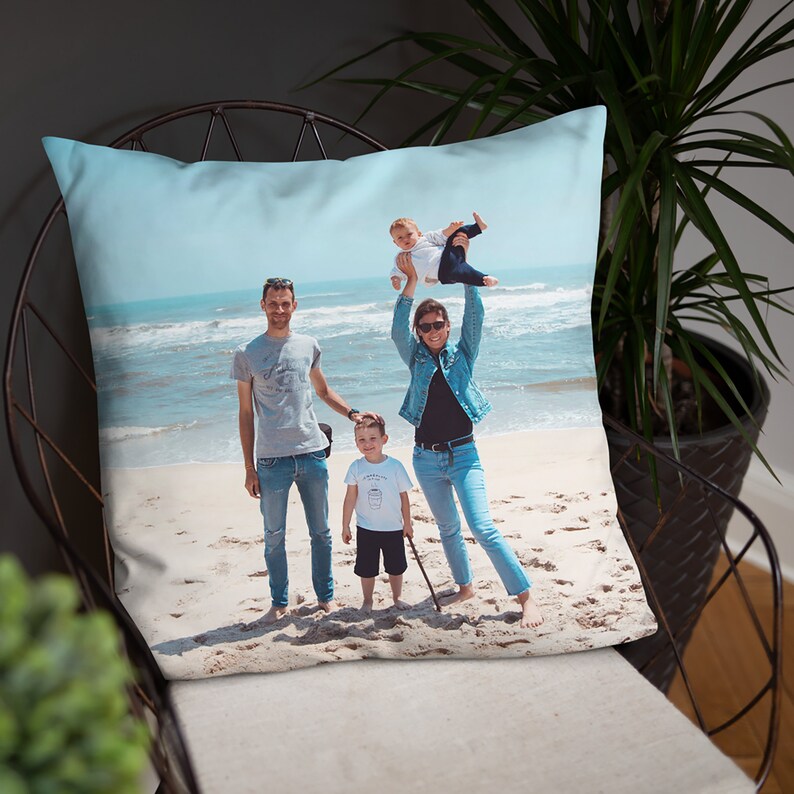 May include: A family of four on a beach. The parents are holding their two children, one in each arm. The family are smiling and looking at the camera. The beach is sandy and the water is blue.