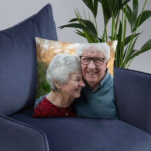 May include: An older couple, a man and a woman, are smiling and looking at each other. The man is wearing a blue shirt and the woman is wearing a red and black patterned shirt. They are standing in front of a blurred background of trees.