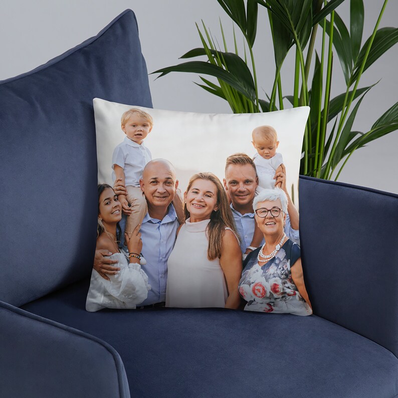 May include: A white cushion featuring a photograph of a family of seven. The family is standing in front of a blurred background. The photo includes two young children, two adults, and three older adults. The adults are smiling and the children are looking at the camera.