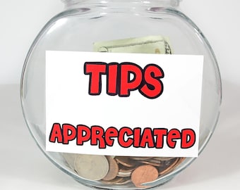 May include: A clear glass fishbowl with a white label that says "TIPS APPRECIATED" in red and black lettering. There are coins and a dollar bill inside the bowl.