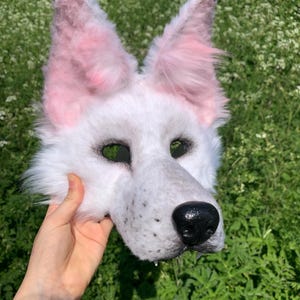 May include: White wolf fursuit head with pink ears and a black nose. The fursuit head is held in a hand.