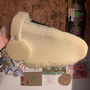May include: A large, cream-coloured foam object, possibly a prop or costume piece, with a rounded shape and a curved section. The object has a smooth texture and appears lightweight. The background includes posters and drawings.