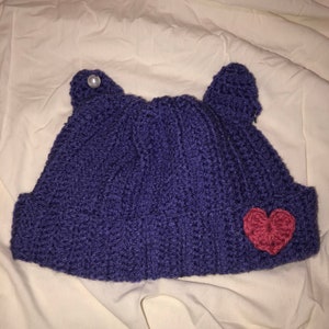Könnte beinhalten: Eine lila gestrickte Mütze mit einem rosa Herz und einem Perlenknopf oben. Die Mütze hat zwei Ohrenklappen.