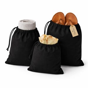 May include: Three black drawstring bags of varying sizes. One bag contains a rolled gray towel, another holds a bar of soap, and the largest bag has a pair of brown leather sandals with a tag that reads "TRAVEL".