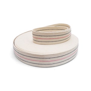 May include: A roll of off-white cotton webbing with black and pink stripes. The webbing is rolled up and the ends are visible.