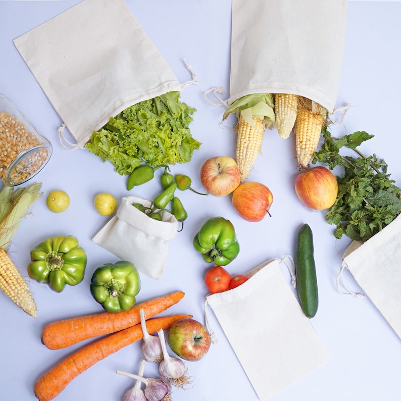 May include: A flat lay of various produce and white cotton bags. The produce includes green peppers, red apples, corn on the cob, green lettuce, and carrots. The bags are made of natural cotton and are perfect for storing and carrying groceries.