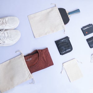 May include: A flat lay image of various items, including two pairs of white sneakers, a red t-shirt, a white drawstring bag, a black wallet, a white drawstring bag with a black zipper, and a white drawstring bag with a wooden paddle.