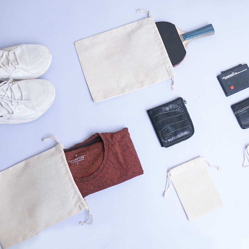 May include: A flat lay image of various items, including two pairs of white sneakers, a red t-shirt, a white drawstring bag, a black wallet, a white drawstring bag with a black zipper, and a white drawstring bag with a wooden paddle.