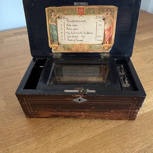 May include: A vintage wooden music box, its lid open to show a list of songs and decorative illustrations. The box is dark brown with a keyhole and a small key. The interior mechanism is visible.